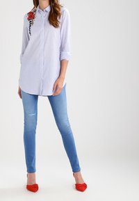 Camisa de botones a rayas azul y blanca con bordado de flores en el hombro izquierdo, combinada con jeans skinny azul claro y zapatos planos rojos.