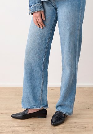 Personne portant un jean bleu clair à jambes larges et des mocassins noirs pointus, debout sur un sol en bois.