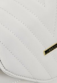 Cartera blanca de cuero acolchado con costura diagonal y una placa de logo en tono dorado. Textura suave y bordes redondeados.