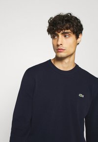 Navy blue long-sleeve sweater made of soft fabric, featuring a small green crocodile logo on the chest and ribbed neckline and cuffs.
