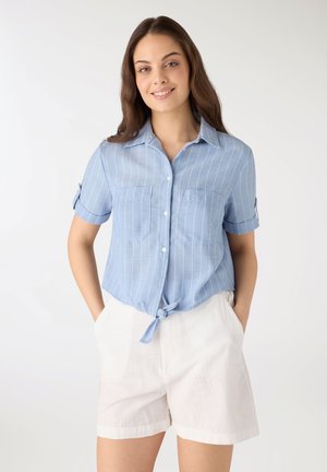 Donna sorridente, indossa una camicia a righe azzurro chiaro a maniche corte annodata in vita e pantaloncini bianchi, in piedi con le mani nelle tasche.