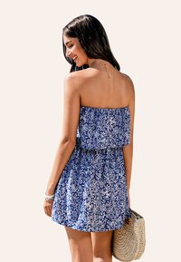 Sukienka bez ramiączek w niebieskim kolorze z białym kwiatowym wzorem, rozkloszowanym dołem i elastyczną talią. Modelka trzyma w plecionej beżowej torbie. Gładka faktura materiału.