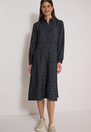 Femme portant une robe midi à manches longues imprimé léopard bleu marine avec col zippé et baskets beiges, debout devant un fond uni.