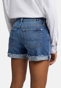Shorts en denim bleu moyen avec un ourlet effiloché, présentant un design classique à cinq poches, des passants de ceinture et une étiquette de marque à l'arrière.