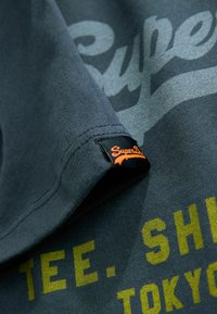 Tmavohnedá bavlnená tričko s žltým textom "TEE. SHIRT TOKYO." Obsahuje značkovú čiernu etiketu s oranžovým textom na leme.