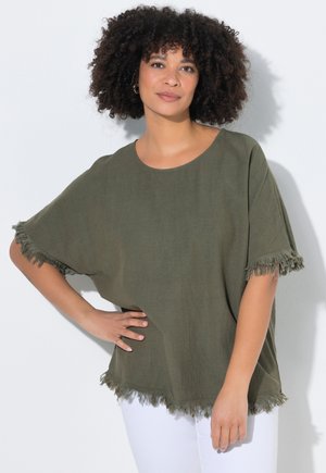 Kvinde med krøllet hår iført en olivengrøn kortærmet top med frynser og hvide bukser, stående med den ene hånd på hoften mod en hvid baggrund.