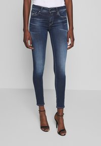Jeans oscuros ajustados de mezclilla, con un acabado desgastado, diseño de cinco bolsillos y costuras en contraste. Combinados con sandalias negras de tacón.