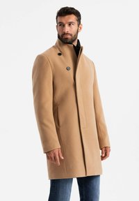 Wollmantel in Camel mit hohem Kragen, zwei schwarzen Knöpfen und einem schlanken Design. Kombiniert mit blauen Jeans für einen rundum gelungenen Look.