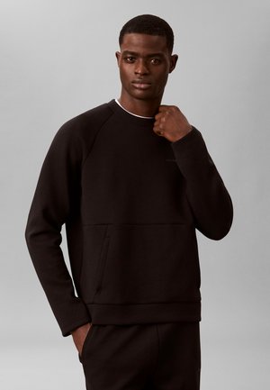 Jeune homme portant un sweat-shirt noir à manches longues et un pantalon assorti, debout devant un fond gris uni.