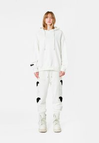Sudadera blanca de gran tamaño con cordones, combinada con pantalones de chándal blancos que tienen patrones de hojas negras y bolsillos laterales, llevada con botas blancas.