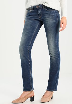 Kvinde iført tætsiddende blå jeans og beige lavblokhælede sko, stående mod hvid baggrund.