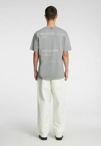 Szary T-shirt z krótkim rękawem z nadrukowanym tekstem o różnych rozmiarach, w zestawieniu z szerokimi białymi spodniami. Na tekście znajdują się napisy "Kopenhaga" i "2025".