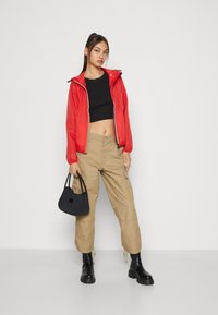 Chaqueta impermeable roja con capucha, top corto negro, pantalones cargo color caqui y botas negras de tobillo. Sosteniendo un pequeño bolso negro.