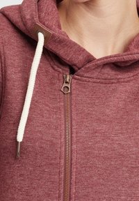 Felpa rossa con zip frontale, dotata di cordino crema e accessori in bronzo. Il materiale è testurizzato con una finitura morbida.