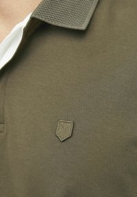 Olivovo zelená polokošela s textúrovaným golierom a malým embosovaným štítom s logom na ľavej strane. Mäkká, hladká tkanina.