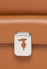 Hnědá kožená kabelka s kovovým zámkem ve tvaru geometrického siluety psa a texturovaným povrchem. Okraje jsou pečlivě prošívané.