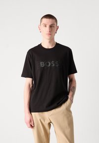Juodas medvilnės marškinėliai su dideliu "BOSS" logotipu priekyje. Trumpomis rankovėmis, apvali iškirptė ir laisvas kirpimas. Dėvimi su smėlio spalvos kelnėmis.
