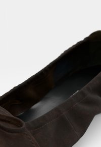 Chaussure plate marron avec un bout rond, faite d'un matériau texturé. Présente un petit bouton décoratif sur le côté et une doublure intérieure lisse.