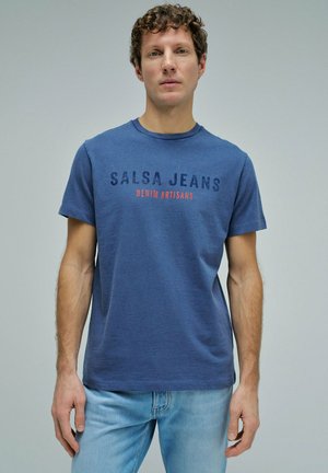 Hombre con una camiseta azul que dice "SALSA JEANS DENIM ARTISANS" y unos vaqueros azul claro, de pie frente a un fondo gris liso.