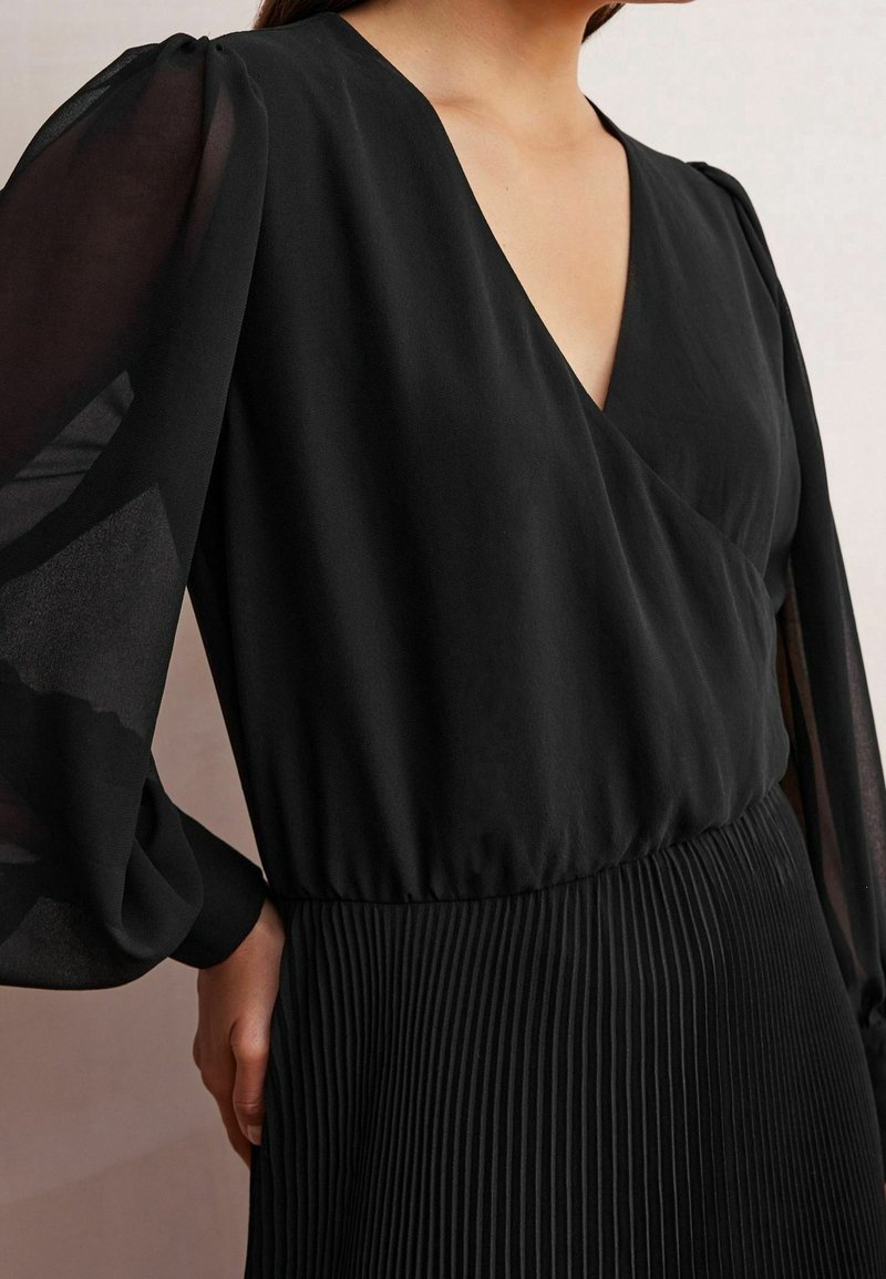 Haut noir avec un décolleté en V profond et de longues manches bouffantes transparentes ; jupe plissée noire à la taille, créant une silhouette fluide.