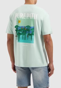 Mintgroene t-shirt met korte mouwen, met een kleurrafische afbeelding van groene stoelen en tafels, met daarboven de tekst "PURE PATH". Zacht katoenmateriaal.