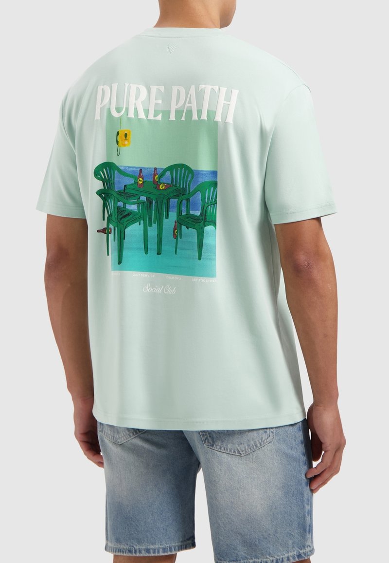 Mintgroene t-shirt met korte mouwen, met een kleurrafische afbeelding van groene stoelen en tafels, met daarboven de tekst "PURE PATH". Zacht katoenmateriaal.