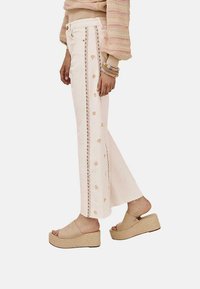 Pantalones de algodón blancos con bordado decorativo rosa a lo largo de los lados. Diseño de pierna ancha, con un dobladillo deshilachado y combinados con sandalias de plataforma marrones.