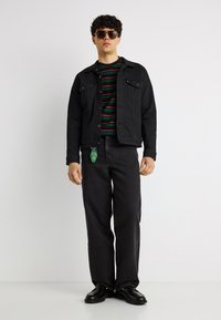 Νεαρός άνδρας με σκούρα γυαλιά ηλίου, μαύρο τζιν μπουφάν και παντελόνι, ριγέ πουκάμισο, μαύρα παπούτσια, στέκεται μπροστά σε λευκό φόντο.