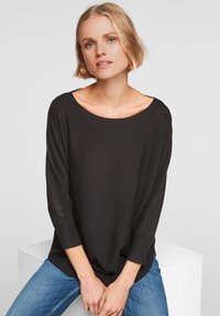 Zwarte langwerpige top met een brede halslijn en een iets losse pasvorm, gecombineerd met blauwe jeans. Zachte textuur, casual ontwerp, geen zichtbare patronen.