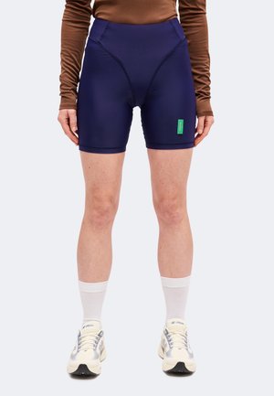 Mannequin portant un short de cyclisme bleu marine avec une étiquette verte, un haut marron à manches longues, des chaussettes blanches et des baskets blanches.
