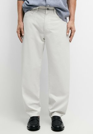 Mann trägt lockere weiße Hose, schwarze Schuhe und ein blau kariertes Hemd, die Hände ruhen seitlich.