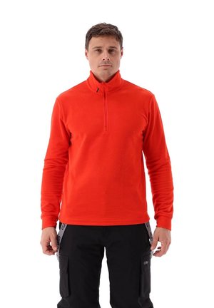 Homme debout portant un pull polaire zippé orange vif et un pantalon cargo noir avec des bretelles, faisant face à un fond blanc.