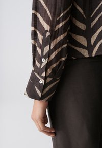 Sleeve of brown patterned blouse with buttoned cuff on extended arm over black skirt or pants against plain background.