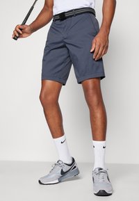Herregrå shorts laget av lett stoff, med flat front og oppbrettede sømmer. Kombinert med svart belte og grå joggesko.