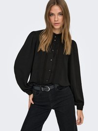 Svart blus med puffärmar, subtil textur och vertikala veck framtill. Kombinerad med mörka jeans och ett grått bälte.