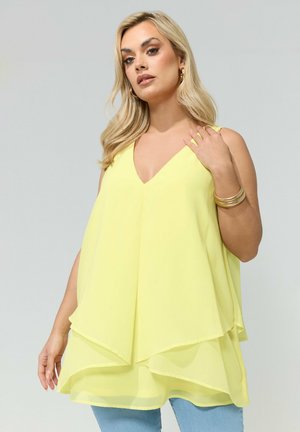 Blondynka w żółtej, warstwowej bluzce bez rękawów, niebieskich dżinsach, złotych kolczykach kołach i złotych bransoletkach na szarym, jednolitym tle.