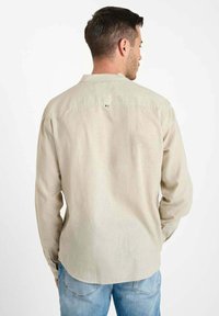 Man in een beige overhemd met lange mouwen en een lichtblauwe spijkerbroek, gezien van achteren tegen een witte achtergrond.