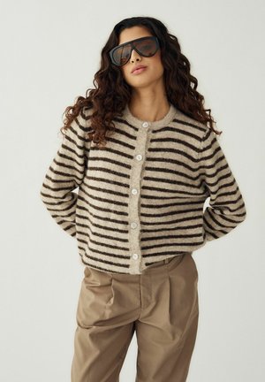 Ženska z dolgimi kodrastimi lasmi, ki nosi velike črne sončna očala, bež in rjavo črtasto kardigan s gumbi ter bež hlače, ob postavljeni proti enostavnem ozadju.