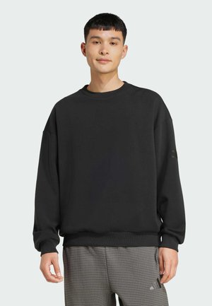 Jeune homme portant un sweat-shirt noir ample et un pantalon gris à carreaux, debout devant un fond clair uni.