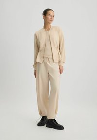 Ensemble d'athlétisme beige comprenant une veste bomber zippée avec une taille cintrée et un pantalon assorti avec un cordon de serrage. Des bottes noires à lacets complètent le look.