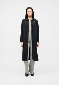 Γυναίκα στέκεται φορώντας ένα μακρύ μαύρο ανοιχτό παλτό πάνω από ένα μπεζ τοπ και ανοιχτό γκρι παντελόνι φαρδιάς γραμμής με μαύρα μυτερά παπούτσια, μπροστά σε ένα απλό φόντο.