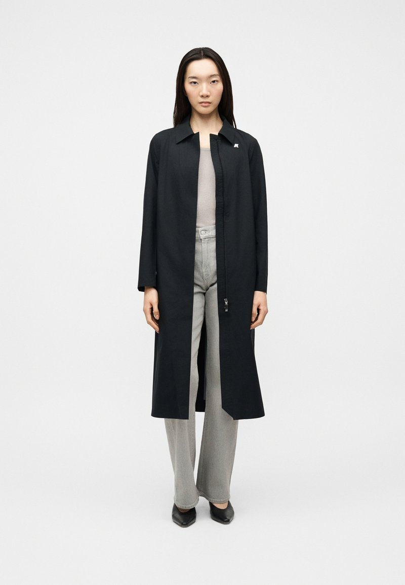 Γυναίκα στέκεται φορώντας ένα μακρύ μαύρο ανοιχτό παλτό πάνω από ένα μπεζ τοπ και ανοιχτό γκρι παντελόνι φαρδιάς γραμμής με μαύρα μυτερά παπούτσια, μπροστά σε ένα απλό φόντο.