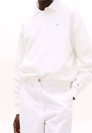 Personne portant une chemise blanche à manches longues avec col et un pantalon blanc, debout avec une main dans la poche sur un fond uni.