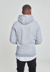 Felpa grigia con cappuccio realizzata in tessuto morbido, con tasca frontale, bordi a coste sui polsini e sull'orlo. Vista posteriore, con una camicia bianca a strati sotto.