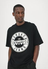 Juodas trumpomis rankovėmis marškinėliai, kuriuose yra apvalus baltas "Versace Couture Jeans" logotipas su metais "1988" centre. Pagaminta iš medvilnės.