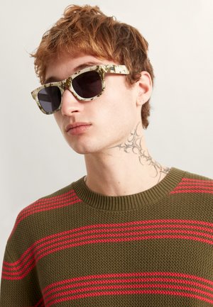 Lunettes de soleil à motif camouflage avec verres foncés, monture carrée et surface texturée. Le modèle porte un pull vert à rayures.