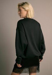 Czarny oversize'owy sweatshirt z opadającymi ramionami, połączony z przejrzystą czarną koronkową spódnicą. Materiał jest miękki, ma luźny krój i widoczne szwy.