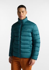 Teal pufferska jakna s horizontalnim prošivenim dizajnom, visokim ovratnikom i prednjim zatvaračem. Ima dvije bočne džepove, napravljena je od laganog materijala.