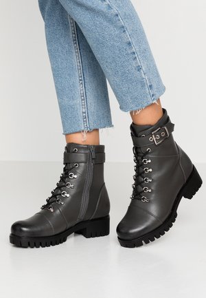 Botas de tobillo de cuero gris con suelas gruesas, herrajes plateados, cordones y una cremallera lateral. Presentan un detalle de hebilla en la parte superior.