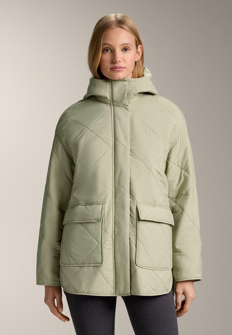 Žena nosí svetlozelenú prešívanú bundu s vysokým golierom, veľkými prednými vreckami a kapucňou, v kombinácii s tmavými legínami, neutrálne pozadie.